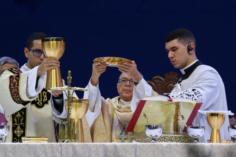 A imagem mostra uma cerimônia religiosa, provavelmente uma missa, onde três homens estão em destaque. Um deles, ao centro, é um sacerdote mais velho, que está vestido com vestes litúrgicas claras e segurando um cálice. À sua esquerda, um homem jovem, também vestido com vestes litúrgicas, levanta um cálice dourado. À direita, outro jovem, vestido de maneira similar, segura uma patena. O fundo é de cor azul escura e a mesa do altar está decorada com toalhas e utensílios litúrgicos.
