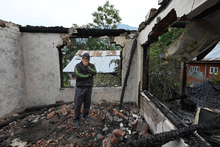 Um homem está em pé dentro de uma casa que foi parcialmente destruída por um incêndio. As paredes estão chamuscadas e há escombros de tijolos no chão. O homem parece estar pensativo e triste, com as mãos no rosto. Ao fundo, é possível ver uma árvore e uma estrutura de telhado de metal.