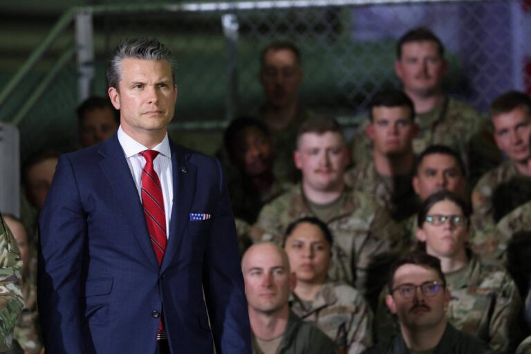 Um homem em traje formal, com um terno escuro e uma gravata vermelha, está em pé em primeiro plano. Ao fundo, há um grupo de militares em uniformes camuflados, alguns com expressões sérias. O ambiente parece ser uma instalação militar, com uma grade ao fundo.