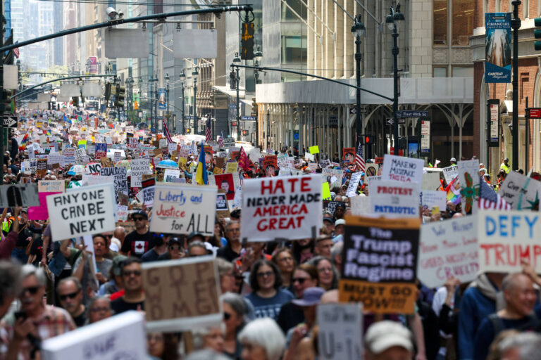 A imagem mostra uma grande multidão participando de um protesto em uma rua urbana. As pessoas seguram diversos cartazes com mensagens de protesto, como
