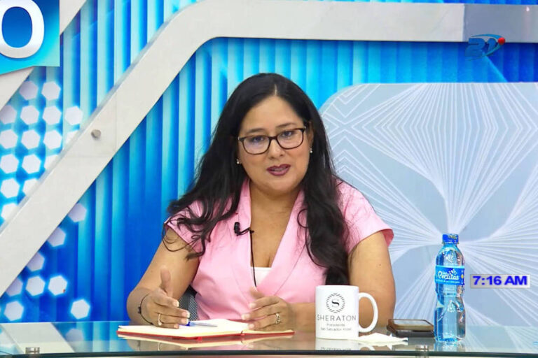 Uma mulher com cabelo longo e liso, usando uma blusa rosa, está sentada em uma mesa de vidro em um estúdio de televisão. Ela usa óculos e parece estar falando ou apresentando algo. Na mesa, há um caderno e uma caneta, além de uma caneca branca com um logotipo. Ao fundo, há um painel azul com elementos gráficos e a hora marcada como 7:16 AM.
