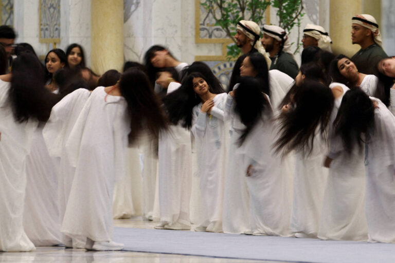 Um grupo de mulheres vestindo longos vestidos brancos está se apresentando em um ambiente interno. Elas estão em movimento, com os cabelos longos balançando, enquanto algumas delas inclinam a cabeça para trás. Ao fundo, há homens com trajes tradicionais, observando a performance. O ambiente é decorado com colunas e plantas, sugerindo um espaço elegante.