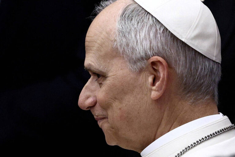 A imagem mostra o perfil de um homem com cabelo grisalho e uma veste branca, usando um chapéu branco. Ele tem uma expressão serena e está voltado para a direita. O fundo é escuro, destacando a figura do homem.