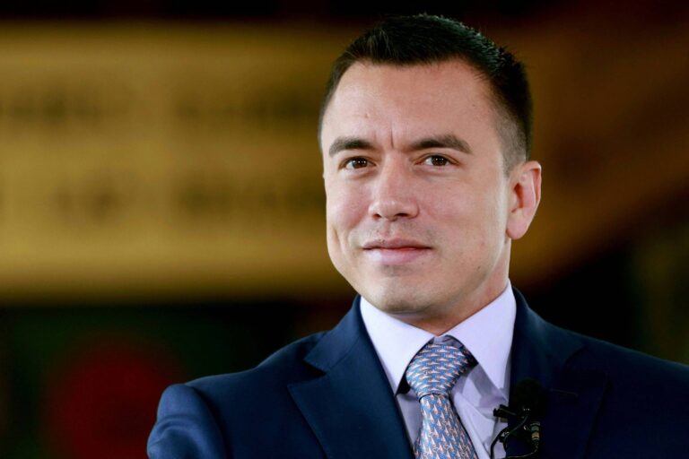 Um homem com cabelo curto e escuro, usando um terno azul e uma gravata com padrão claro. Ele está sorrindo levemente e olhando para a câmera. O fundo é desfocado, mas parece ter elementos de um ambiente formal.