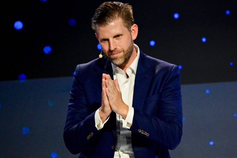 Um homem está em um palco, vestindo um terno azul escuro e uma camisa branca. Ele está com as mãos juntas em um gesto de oração ou agradecimento, olhando para a frente. O fundo é escuro com pontos de luz azul, criando um efeito visual interessante.