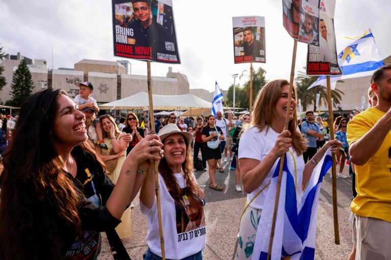 A imagem mostra um grupo de pessoas participando de um protesto. Algumas mulheres estão na frente, segurando cartazes que pedem para trazer alguém de volta para casa. Elas estão sorrindo e vestindo camisetas com imagens. Ao fundo, há uma multidão e bandeiras de Israel. O ambiente parece ser ao ar livre, com árvores e um edifício visível ao fundo.