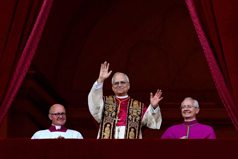 A imagem mostra três homens em trajes religiosos, com um deles no centro acenando. O homem central usa uma vestimenta elaborada, enquanto os outros dois estão ao seu lado, um com uma camisa branca e o outro com uma vestimenta roxa. Eles estão em uma varanda com cortinas vermelhas ao fundo.