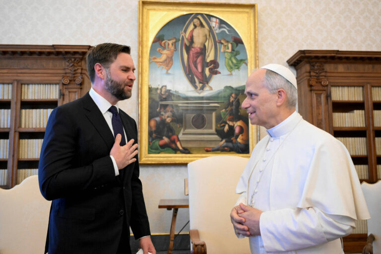 A imagem mostra um encontro entre duas pessoas em um ambiente formal. À esquerda, um homem de terno escuro, com cabelo curto e barba, está em uma posição de respeito, com a mão sobre o coração. À direita, um homem vestido com vestes brancas, que parece ser uma figura religiosa, está sorrindo e olhando para o outro. Ao fundo, há uma pintura clássica que retrata uma cena religiosa, com detalhes em cores vibrantes. O ambiente é decorado com estantes de livros e uma parede com padrão claro.