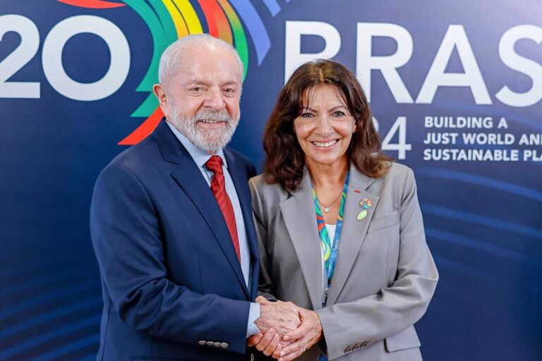 Na imagem, duas pessoas estão se cumprimentando com um aperto de mão. À esquerda, um homem de cabelo grisalho e barba branca, vestido com um terno escuro e uma gravata vermelha. À direita, uma mulher com cabelo castanho e liso, usando um blazer cinza e uma blusa colorida. Ao fundo, há um painel com as palavras