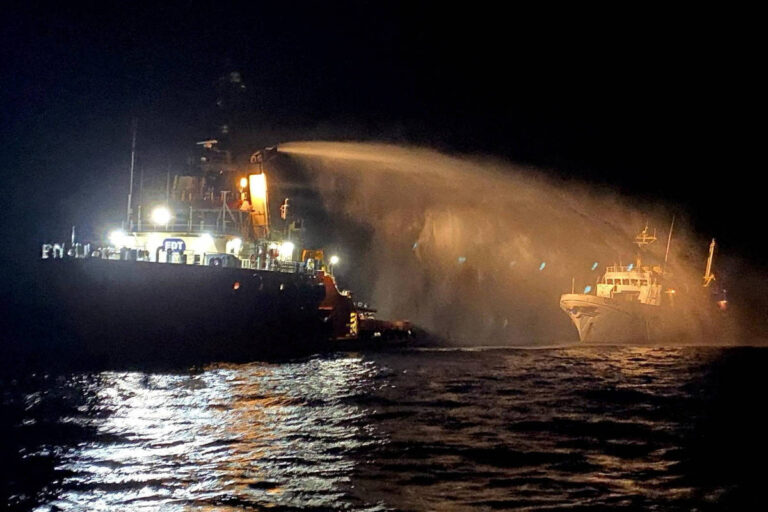 A imagem mostra dois barcos no mar à noite. Um dos barcos, à esquerda, está equipado com luzes brilhantes e está lançando água, possivelmente em uma operação de resgate ou combate a incêndio. O outro barco, à direita, parece ser menor e está próximo ao primeiro. A superfície da água reflete as luzes dos barcos, criando um efeito luminoso.