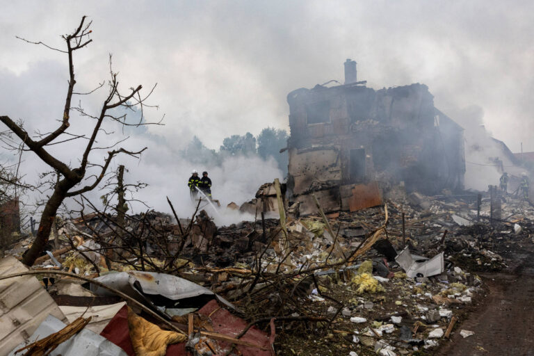 A imagem mostra os destroços de uma construção em uma área urbana, com fumaça densa no ar. Dois bombeiros estão visíveis, trabalhando entre os escombros. O cenário é de destruição, com árvores secas e lixo espalhado pelo chão.