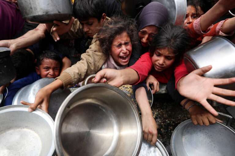 A imagem mostra um grupo de crianças e adultos em uma situação de desespero, tentando pegar panelas durante uma distribuição de alimentos. As expressões faciais refletem angústia e urgência, com algumas pessoas gritando e outras estendendo as mãos em busca de ajuda. O ambiente parece caótico, com várias panelas de metal visíveis.