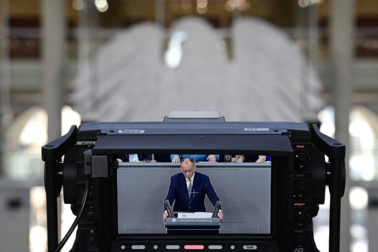 A imagem mostra uma câmera de vídeo focada em um homem que está fazendo um discurso em um palco. O homem está vestido com um terno escuro e está de pé atrás de um púlpito. Ao fundo, é possível ver uma grande estrutura com formato de asas, que parece ser uma instalação artística ou decorativa. A cena sugere um ambiente formal, possivelmente em um evento político ou institucional.