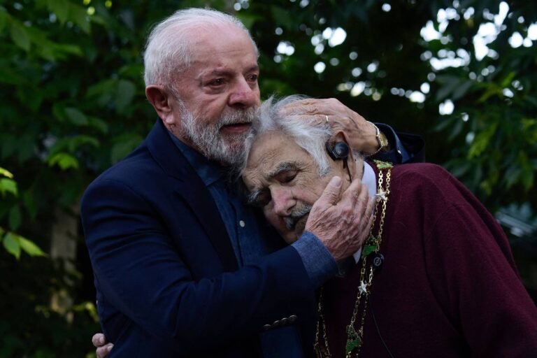 Na imagem, duas pessoas idosas estão se abraçando em um ambiente ao ar livre, cercado por folhagens verdes. A pessoa à esquerda, com cabelo grisalho e barba, veste um paletó escuro e parece estar confortando a outra, que está à direita, com cabelo claro e um suéter marrom. A expressão de ambos transmite um momento de afeto e conexão emocional.