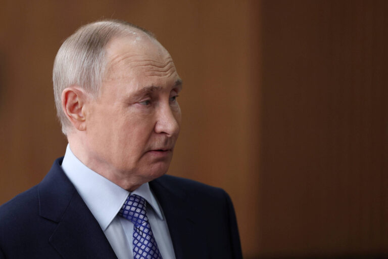 Putin, um homem com cabelo curto e liso, usa um terno escuro e uma camisa clara com uma gravata azul. Ele está olhando para o lado, com uma expressão séria. O fundo é desfocado, sugerindo um ambiente interno.