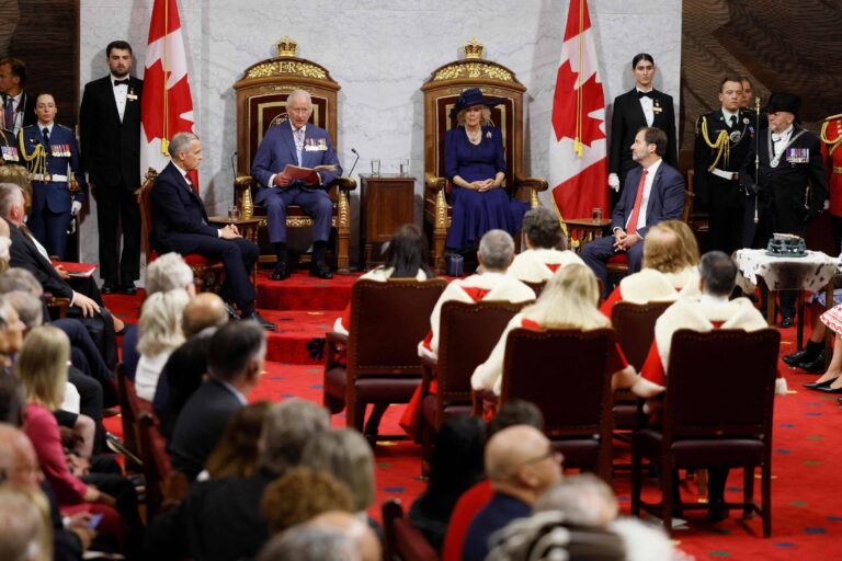 A imagem mostra uma cerimônia oficial no Canadá, com várias pessoas presentes. No centro, há duas cadeiras grandes e ornamentadas, ocupadas por figuras importantes, enquanto outras pessoas estão sentadas em cadeiras ao redor. Bandeiras canadenses estão visíveis ao fundo, e há membros das forças armadas em uniforme. O ambiente é formal e decorado.