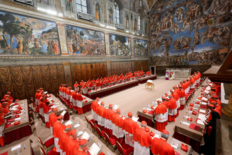 A imagem mostra um conclave no Vaticano, com cardeais vestidos de vermelho reunidos em uma sala. Eles estão dispostos em mesas em forma de U, com um altar ao centro. As paredes são decoradas com afrescos, incluindo obras de arte renascentista. A atmosfera é solene e formal, refletindo a importância do evento.