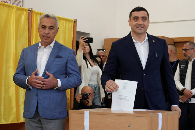 A imagem mostra dois homens em um local de votação. O homem à esquerda, com cabelo grisalho e vestindo um paletó azul claro, está em pé ao lado de um homem à direita, que está segurando um papel e sorrindo. O homem à direita está vestido com um paletó escuro. Ao fundo, há uma cortina amarela e várias pessoas observando a cena, algumas com celulares. Uma caixa de votação está visível à frente dos homens.