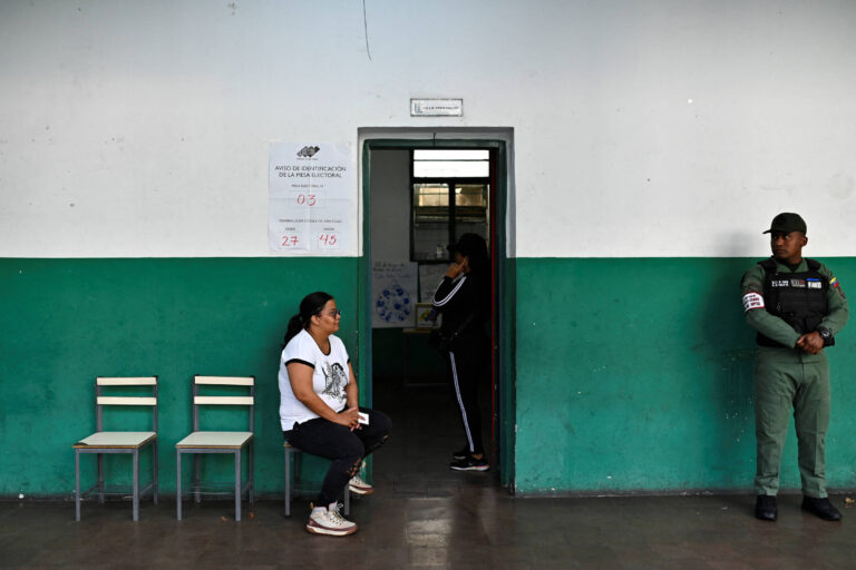 A imagem mostra uma sala de espera com paredes pintadas de verde e branco. À esquerda, há três cadeiras vazias. Uma mulher sentada em uma das cadeiras está vestindo uma camiseta branca e calças escuras. À direita, uma segunda mulher está em pé, parcialmente visível, dentro de uma sala com uma porta aberta. Um homem vestido com uniforme de segurança está em pé ao lado da porta, observando a cena. O ambiente parece ser um espaço público, possivelmente uma instituição.