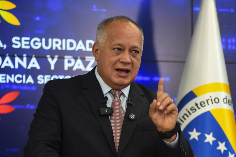Um homem em um terno escuro está falando em um microfone, gesticulando com a mão. Ao fundo, há um painel com texto em espanhol que menciona segurança e paz, e uma bandeira do Brasil é visível à direita.