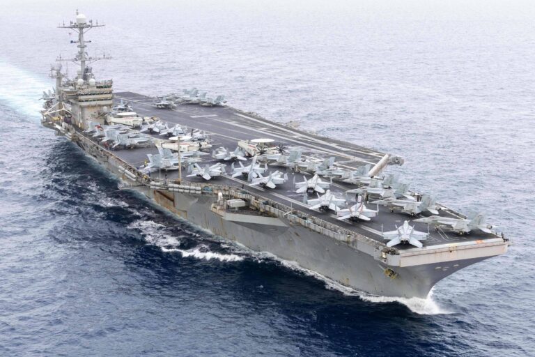 A imagem mostra um porta-aviões navegando em águas abertas. O navio possui uma pista de decolagem visível na parte superior, onde estão estacionados vários aviões de combate. O céu está nublado e o mar parece calmo, com pequenas ondas.