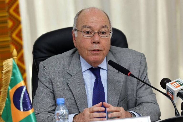 Um homem de óculos, com cabelo grisalho e uma expressão séria, está sentado em uma mesa de conferência. Ele usa um terno cinza e uma gravata azul. À sua frente, há uma garrafa de água e um microfone. Ao fundo, uma bandeira do Brasil está visível, junto a um pano decorativo colorido.