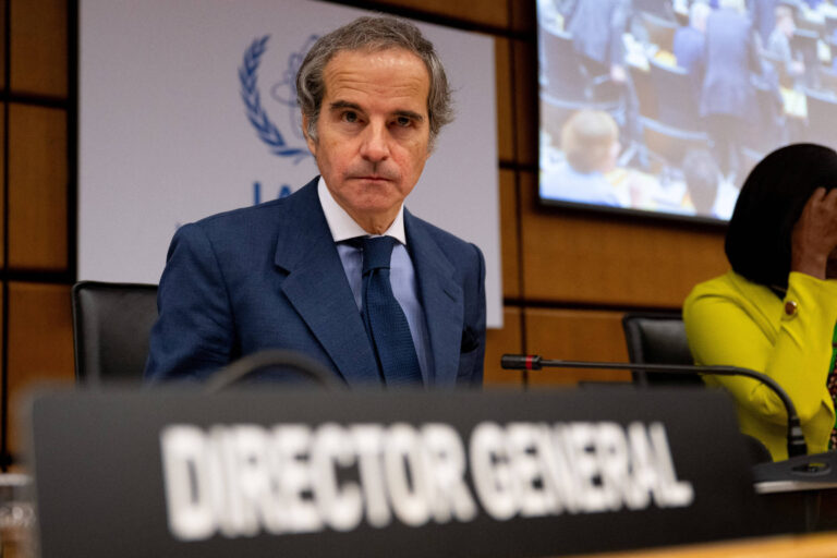 Um homem com cabelo grisalho e terno azul está sentado em uma mesa em uma reunião. Ele parece sério e está olhando para a frente. Na mesa, há uma placa com a inscrição