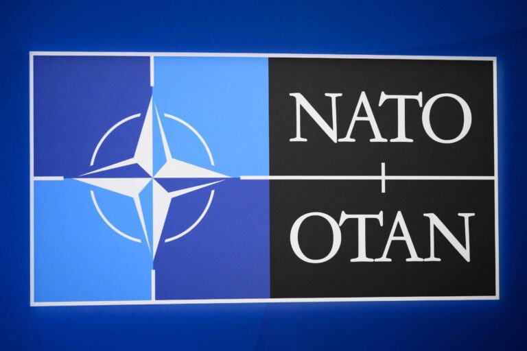 A imagem apresenta o logotipo da OTAN (Organização do Tratado do Atlântico Norte), que é dividido em duas partes. A parte esquerda é azul claro com um símbolo de uma estrela de quatro pontas, enquanto a parte direita é preta com as palavras