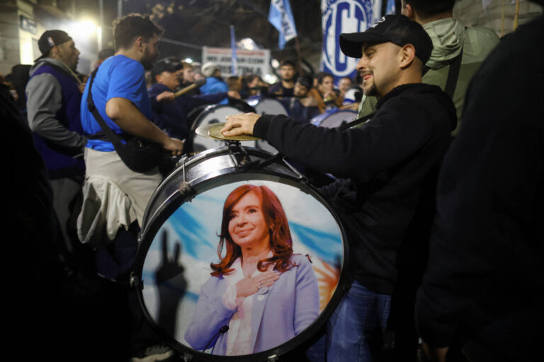 A imagem mostra um grupo de pessoas em uma manifestação. Um homem à direita está segurando um tambor que tem uma foto de uma mulher com cabelo longo e castanho, vestindo um paletó claro. Ao fundo, outras pessoas estão reunidas, algumas segurando instrumentos musicais e bandeiras. A atmosfera parece ser de celebração ou apoio.