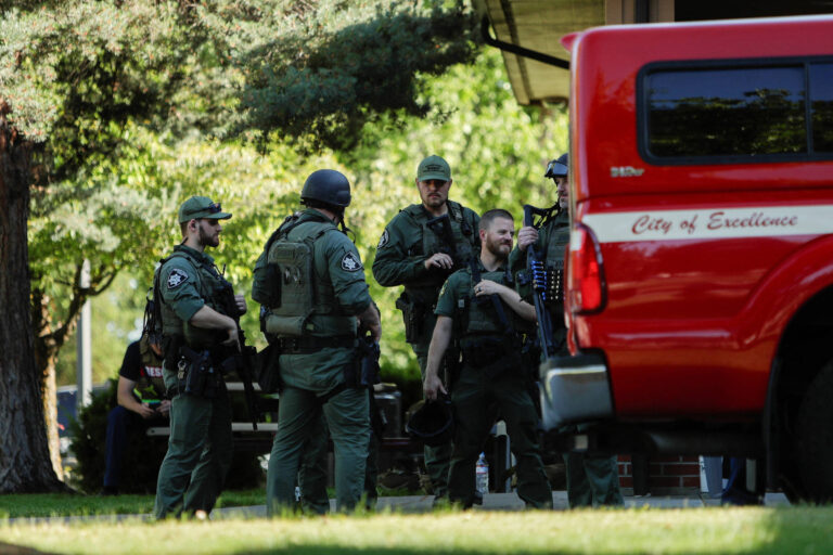 A imagem mostra um grupo de policiais armados e equipados em um ambiente externo, possivelmente em uma operação de segurança. Eles estão vestidos com uniformes táticos e alguns usam capacetes. Ao fundo, há uma caminhonete vermelha com a inscrição