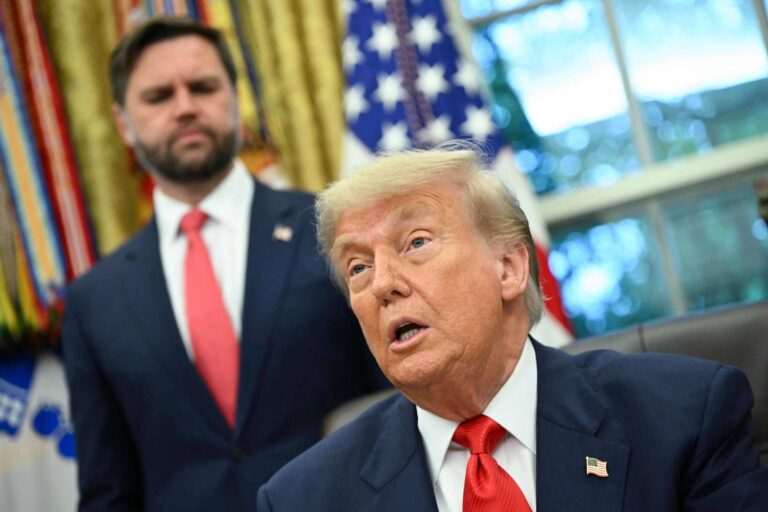 A imagem mostra Donald Trump, um homem de cabelo loiro e terno escuro, falando em um ambiente que parece ser o Salão Oval da Casa Branca. Ao fundo, há J.D. Vance, um homem com cabelo escuro, usando um terno escuro e gravata vermelha, que observa. O ambiente é decorado com cortinas e bandeiras dos Estados Unidos.