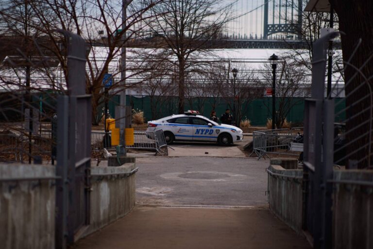 A imagem mostra um veículo da polícia identificado como NYPD estacionado em uma área de parque. Ao fundo, há árvores sem folhas e uma estrutura que parece ser uma ponte. O ambiente é urbano e há algumas pessoas visíveis próximas ao carro da polícia.