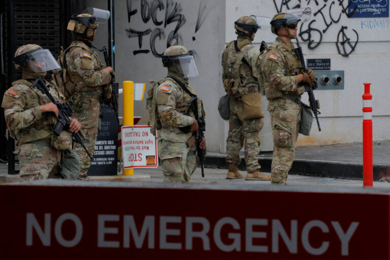 A imagem mostra um grupo de soldados armados, vestidos com uniformes camuflados e equipamentos de proteção, em uma área urbana. Eles estão posicionados em frente a um edifício com grafites nas paredes. Um sinal em primeiro plano indica