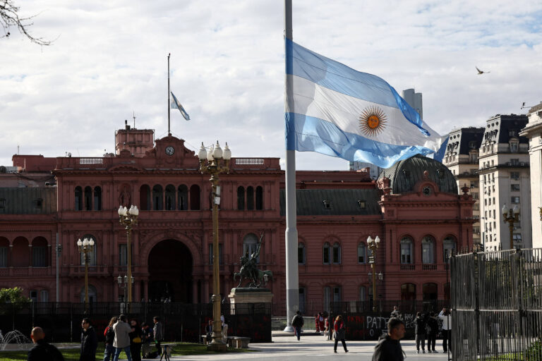 A imagem mostra a Casa Rosada, sede do governo argentino, com uma grande bandeira da Argentina hasteada em um mastro. O edifício é de cor rosa e possui uma arquitetura imponente. No primeiro plano, há pessoas caminhando e o céu está parcialmente nublado.