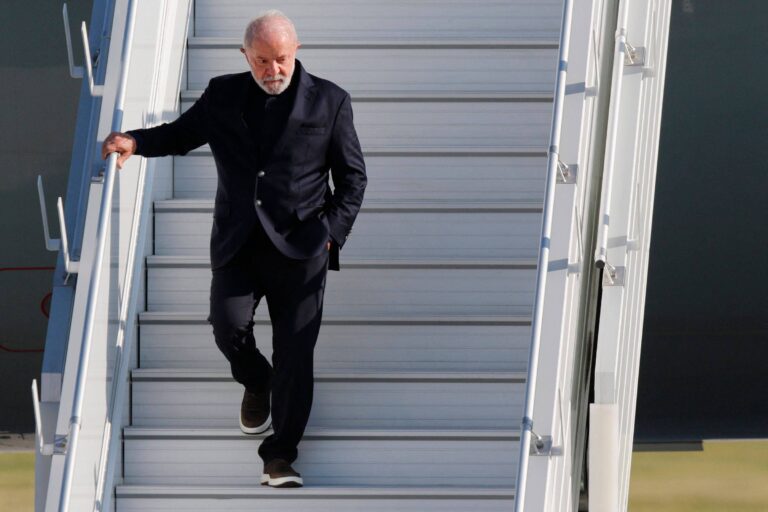 Um homem de cabelos grisalhos e barba, vestido com um terno escuro e sapatos brancos, está descendo as escadas de um avião. Ele parece estar em um aeroporto, com uma expressão séria enquanto caminha.