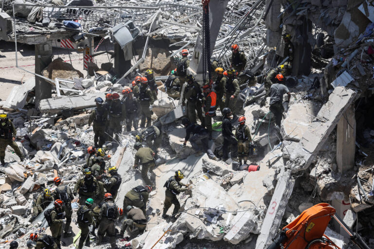 A imagem mostra um grande grupo de socorristas trabalhando em meio aos destroços de um edifício colapsado. Os socorristas estão usando capacetes e equipamentos de segurança, e alguns estão em ação, enquanto outros observam ou se preparam para ajudar. O cenário é caótico, com escombros e estruturas danificadas ao redor.