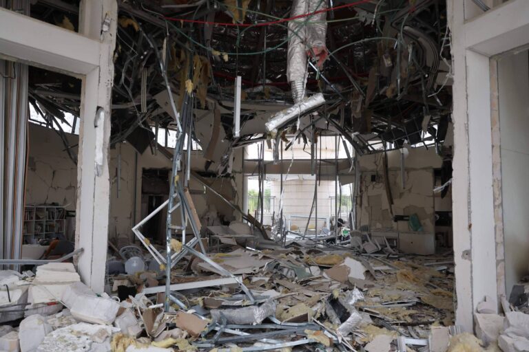 A imagem mostra o interior de um edifício severamente danificado, com paredes destruídas, fiação exposta e detritos espalhados pelo chão. O teto está parcialmente desabado, e há materiais de construção, como pedaços de drywall e tubos, visíveis na cena. A luz entra através de aberturas nas paredes, iluminando a desordem.