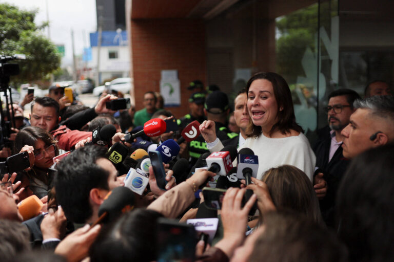 A imagem mostra uma coletiva de imprensa com uma mulher em destaque, falando para um grupo de repórteres. Ela está cercada por jornalistas que seguram microfones e câmeras, enquanto alguns policiais estão ao fundo. A mulher parece estar expressando emoções intensas, e o ambiente é urbano, com prédios visíveis ao fundo.