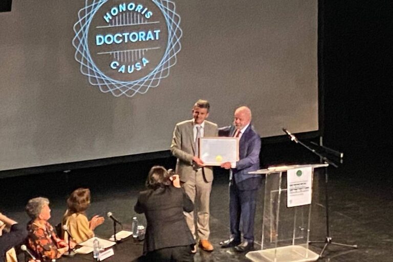 A imagem mostra uma cerimônia onde duas pessoas estão no palco. Uma delas, à esquerda, está segurando um diploma e a outra, à direita, está recebendo o diploma. Ao fundo, há um logotipo que diz