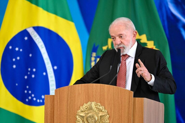 Um homem com cabelo grisalho e barba, vestido com um terno preto e uma gravata vermelha, está falando em um púlpito de madeira. Ao fundo, há bandeiras do Brasil e outras bandeiras em cores verde e azul. O homem parece estar enfatizando um ponto com o dedo levantado.
