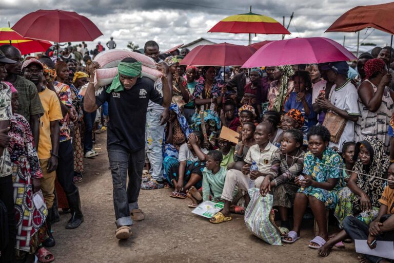 A imagem mostra uma cena de distribuição de alimentos em uma comunidade. Um homem, usando uma camiseta preta e um lenço verde na cabeça, carrega sacos de alimentos nas costas. Ele está cercado por uma multidão de pessoas, muitas das quais seguram guarda-chuvas coloridos. As pessoas na multidão parecem estar atentas e algumas estão sentadas no chão, enquanto outras estão em pé. O ambiente é ao ar livre, com um céu nublado ao fundo.