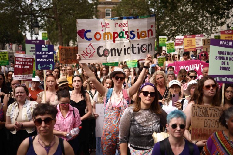A imagem mostra uma grande multidão de mulheres participando de um protesto. Muitas delas seguram cartazes com mensagens sobre direitos reprodutivos, incluindo um que diz