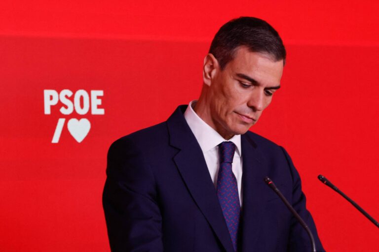 Pedro Sanchez, um homem em um terno escuro está de pé, está olhando para baixo, em um cenário com fundo vermelho. À esquerda, há um logotipo do PSOE, que é um partido político, com um coração ao lado. O homem parece pensativo e sério, com cabelo curto e bem arrumado.