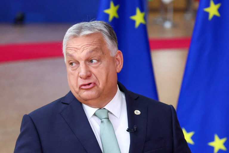 Um homem de cabelo grisalho e pele clara, vestido com um terno escuro e uma gravata clara, está falando em um evento. Ao fundo, há bandeiras da União Europeia com estrelas amarelas em um fundo azul. O homem parece estar expressando uma opinião ou fazendo uma declaração.
