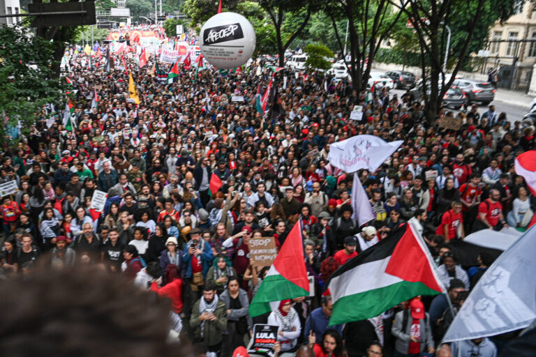 A imagem mostra uma grande multidão reunida em uma manifestação, com pessoas segurando bandeiras e cartazes. Há bandeiras da Palestina e outras representando diferentes grupos. O ambiente é urbano, com árvores ao redor e um balão flutuante com a palavra