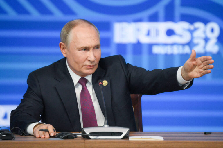 Um homem de terno escuro e gravata roxa está sentado em uma mesa, gesticulando com a mão direita. Ele parece estar falando durante uma conferência de imprensa. Ao fundo, há um painel azul com o texto