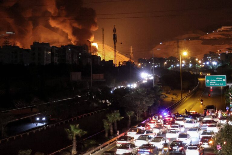 A imagem mostra uma cena noturna de uma área urbana com um incêndio visível ao fundo, emitindo fumaça escura. À frente, há um tráfego intenso de veículos, com muitos carros parados e alguns com luzes acesas. À direita, há uma placa de sinalização indicando direções. O céu está escuro, e a iluminação da cidade é visível, contrastando com a fumaça do incêndio.