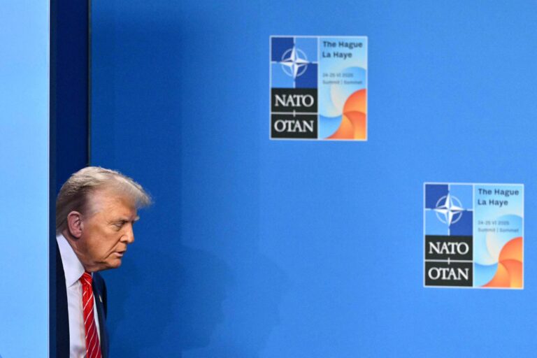Trump, um homem com cabelo loiro e terno escuro, está saindo de um espaço delimitado por uma parede azul. Ele parece estar se aproximando de um evento, com cartazes visíveis ao fundo que mencionam a OTAN e a cidade de Haia. Os cartazes têm o logotipo da OTAN e estão em um fundo azul.