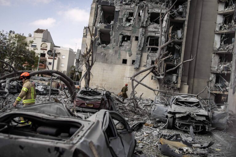 A imagem mostra um cenário de destruição em uma área urbana, com um edifício gravemente danificado, apresentando paredes desmoronadas e janelas quebradas. No primeiro plano, há vários carros danificados e amassados, cobertos por destroços. Um bombeiro, vestido com uniforme e capacete, caminha entre os escombros, enquanto outra pessoa é vista ao fundo, também em meio à destruição. O céu está claro, sugerindo que a cena foi capturada durante o dia.