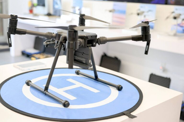 Um drone preto está posicionado em cima de um círculo azul com um símbolo de pouso. O drone possui quatro hélices e uma estrutura robusta, com uma câmera visível na parte frontal. Ao fundo, há uma parede com várias imagens e equipamentos relacionados a drones.